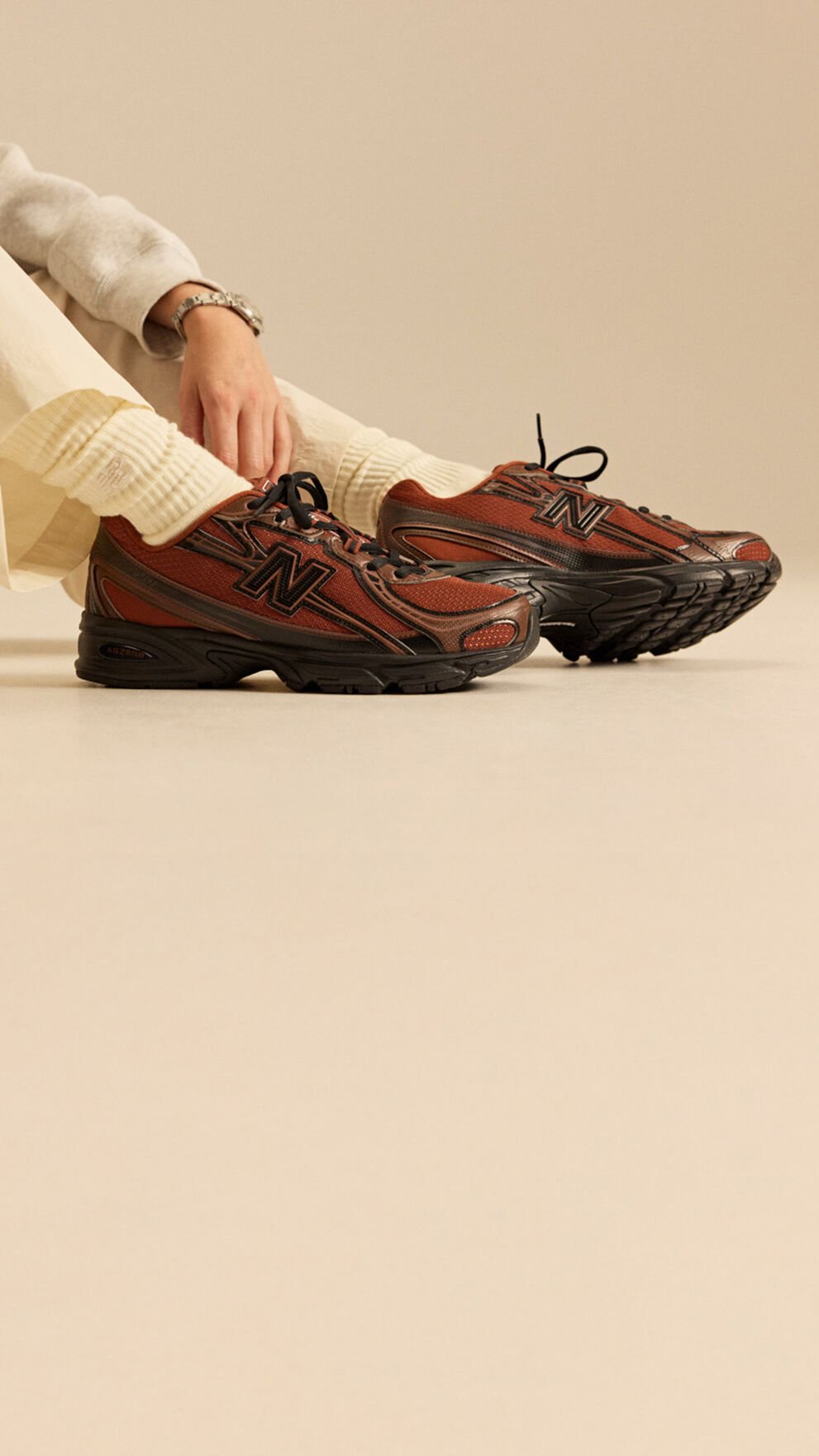 mage of someone sitting wearing New Balance sneakers in front of a beige background. 